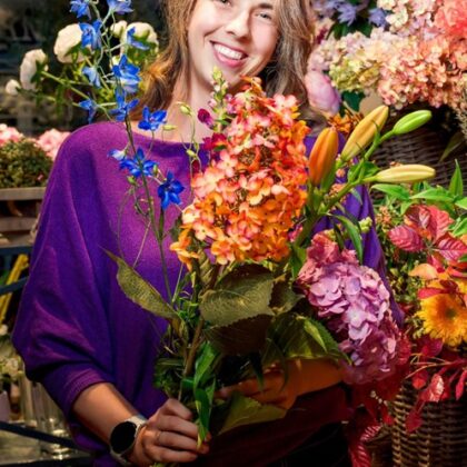 Smiling woman in a purple top holding a large, colorful bouquet in a flower shop.