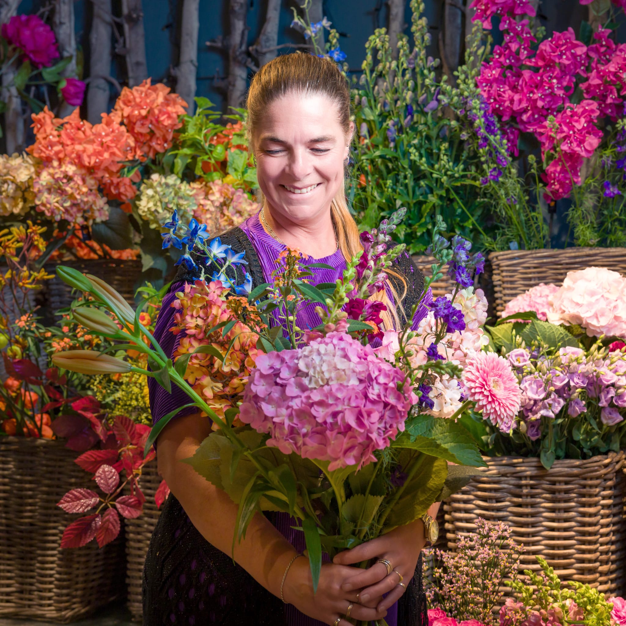 Smiling woman holding a vibrant bouquet of pink, purple, and orange flowers in a flower stall backdrop.