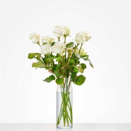 White roses arranged in a tall clear vase with water on a white surface, showing green stems and leaves.