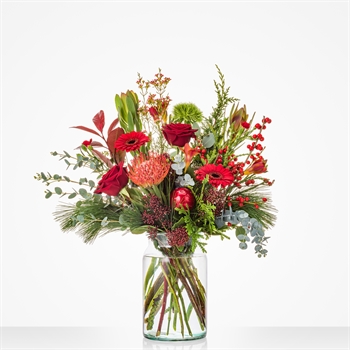 Bouquet of red, pink, and orange flowers with greenery in a tall clear glass vase on a white background.