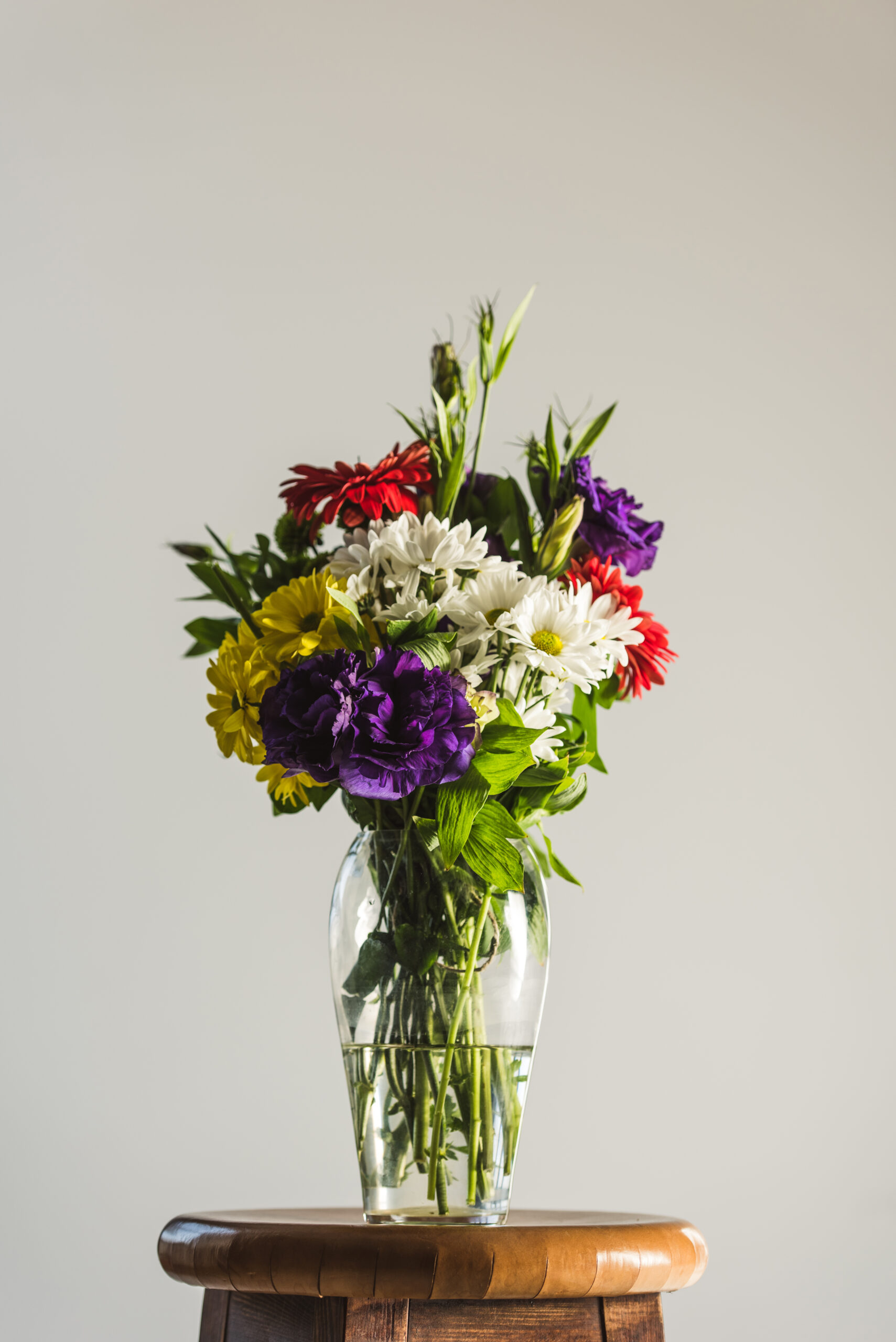 Colorful bouquet of white, yellow, red, purple daisies in a clear glass vase on a wooden stool against a light background.