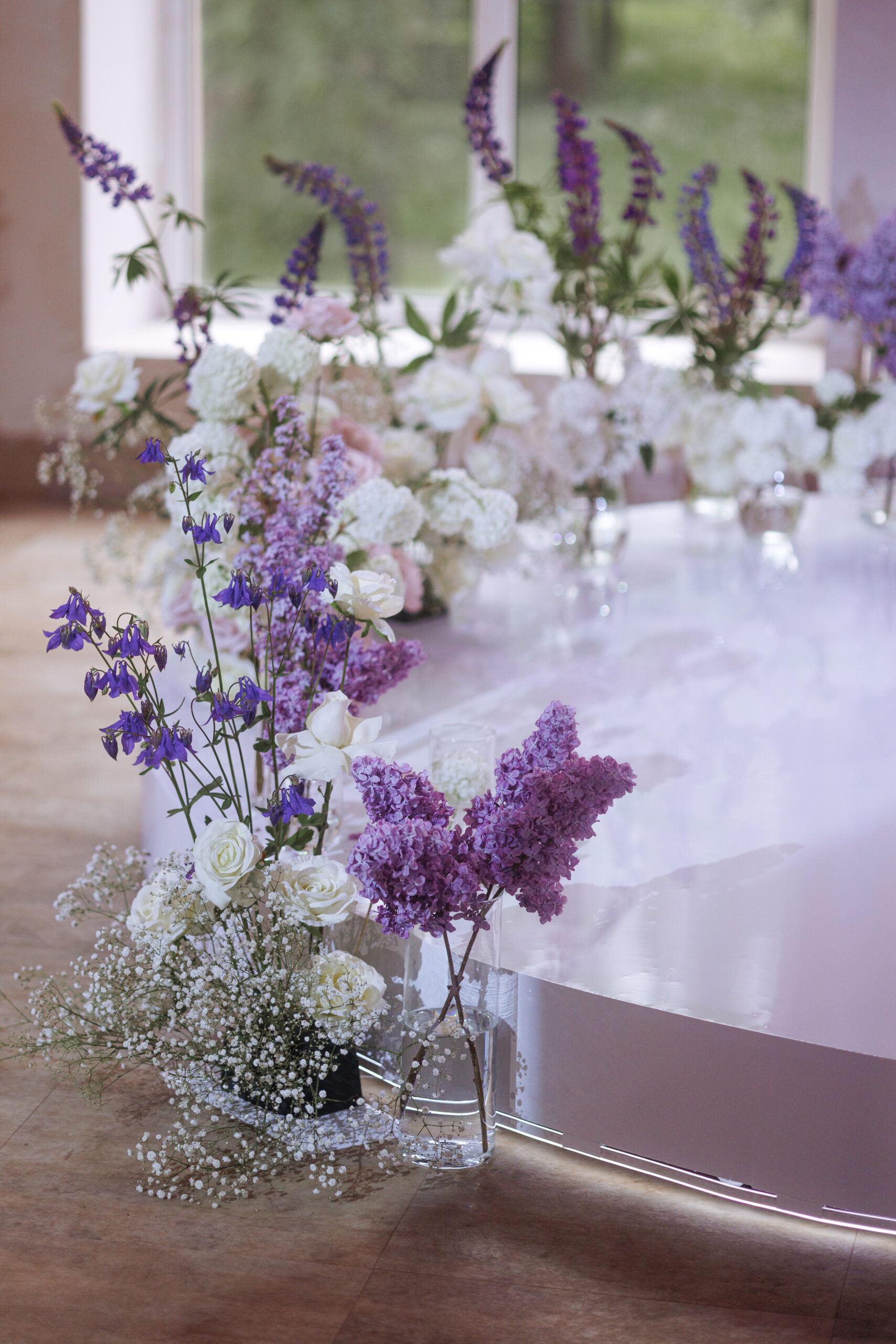 Elegant wedding table centerpiece with white roses, purple lilacs, and baby's breath along a glossy curved table edge.